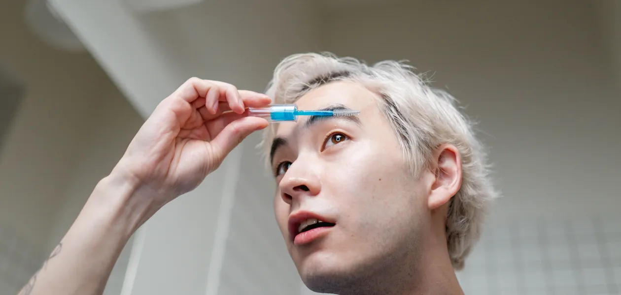 young man grooming his eyebrows with a spoolie brush in a bathroom mirror