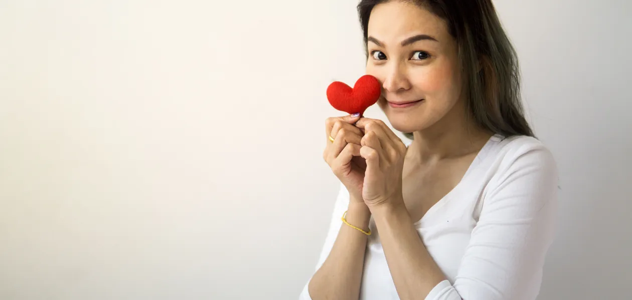 Cheerful female with long hair looking at camera while standing on white background with red heart near face in room