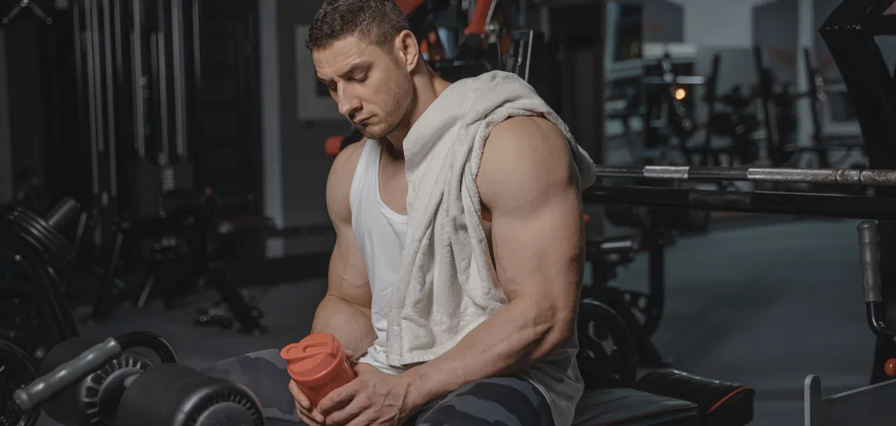 Muscular man sits in gym holding protein shaker after a workout, towel on shoulder.